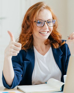 Lächelnde Frau mit Brille am Schreibtisch zeigt den Daumen nach oben, weil ihr eigener Onlinekurs fertig ist